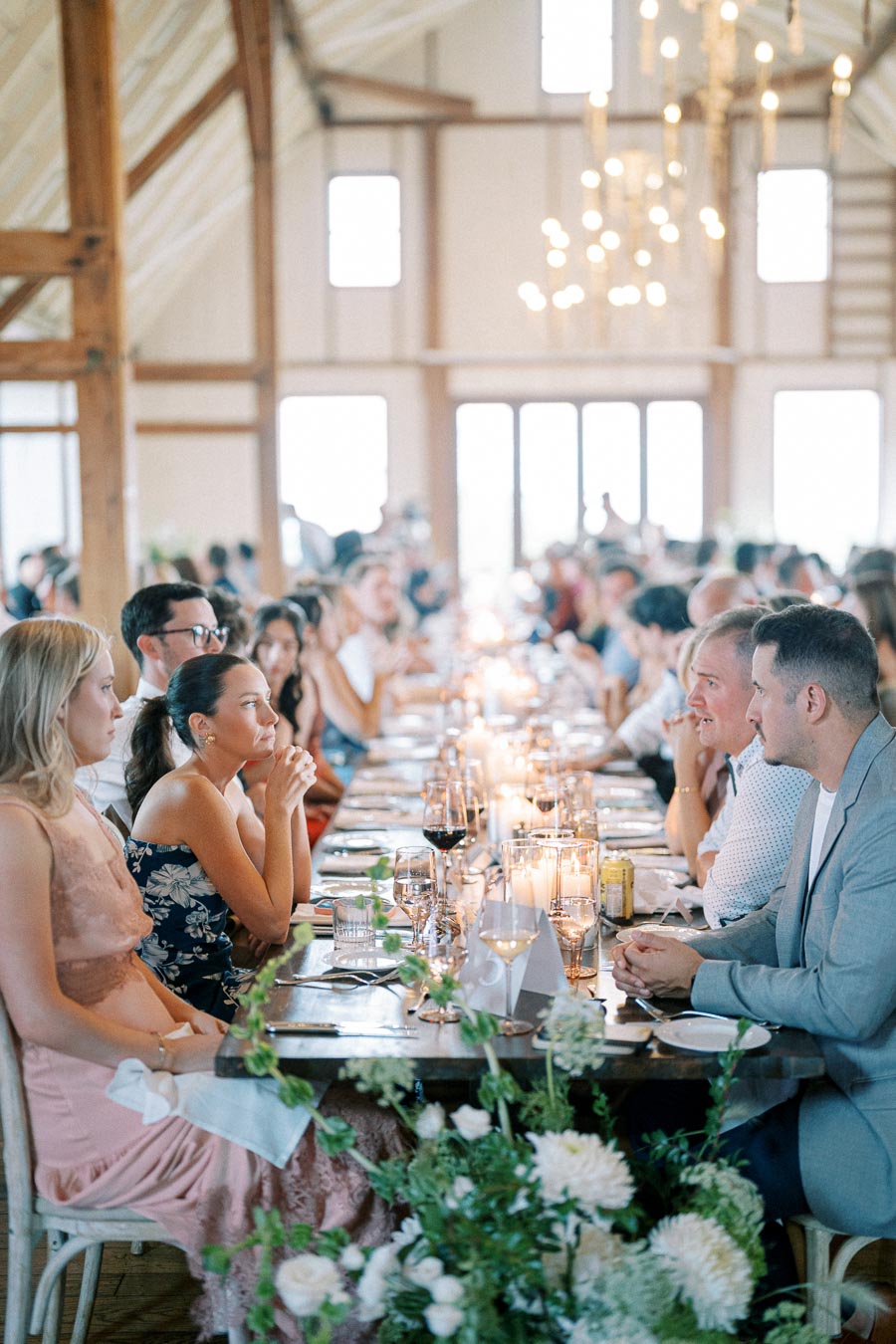 Elegant wedding reception in a rustic barn setting, guests seated at a long table with floral decorations and candlelit ambiance.