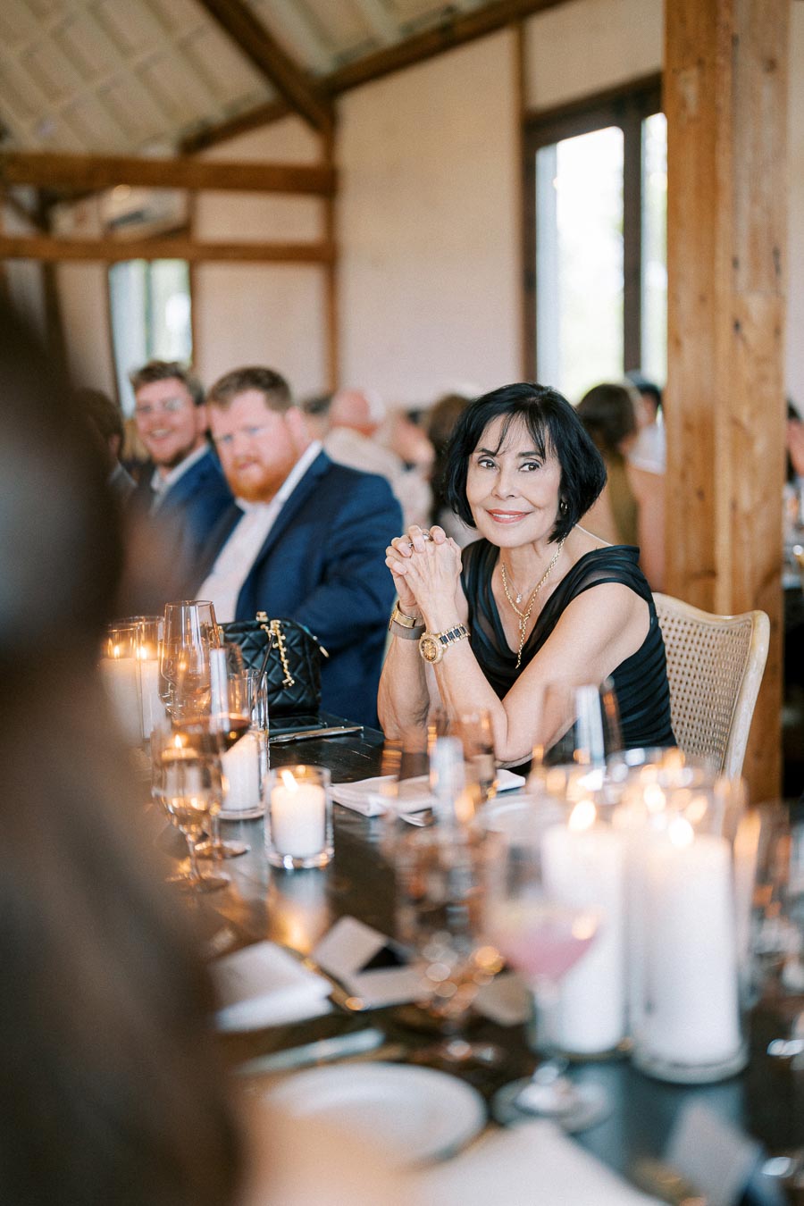 Guests enjoying a formal dinner at a beautifully lit event, featuring elegantly set tables with candles and glassware in a rustic venue.