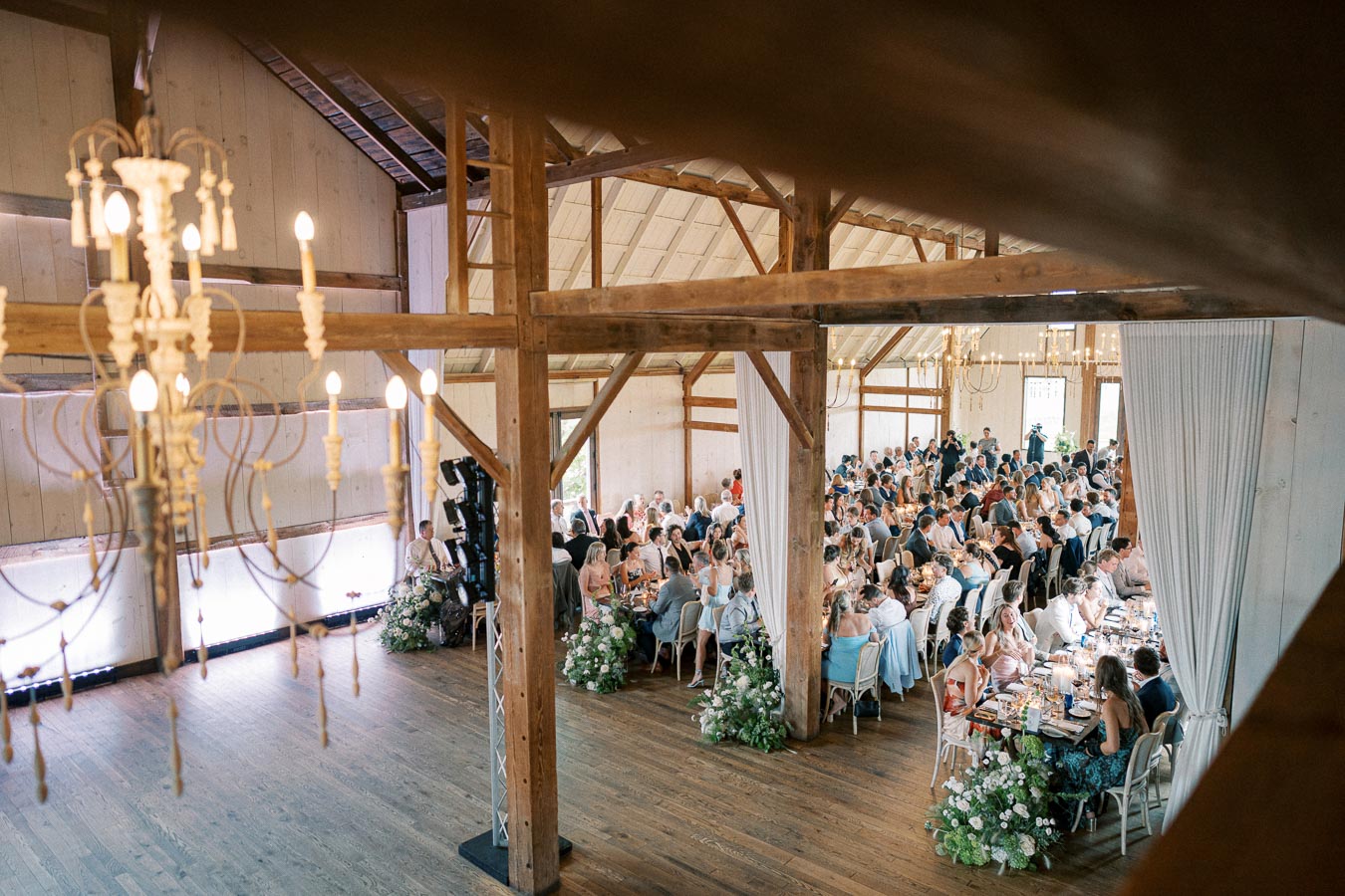 Rustic wedding reception in a spacious barn with elegant chandeliers, featuring guests seated at decorated tables adorned with greenery and flowers.