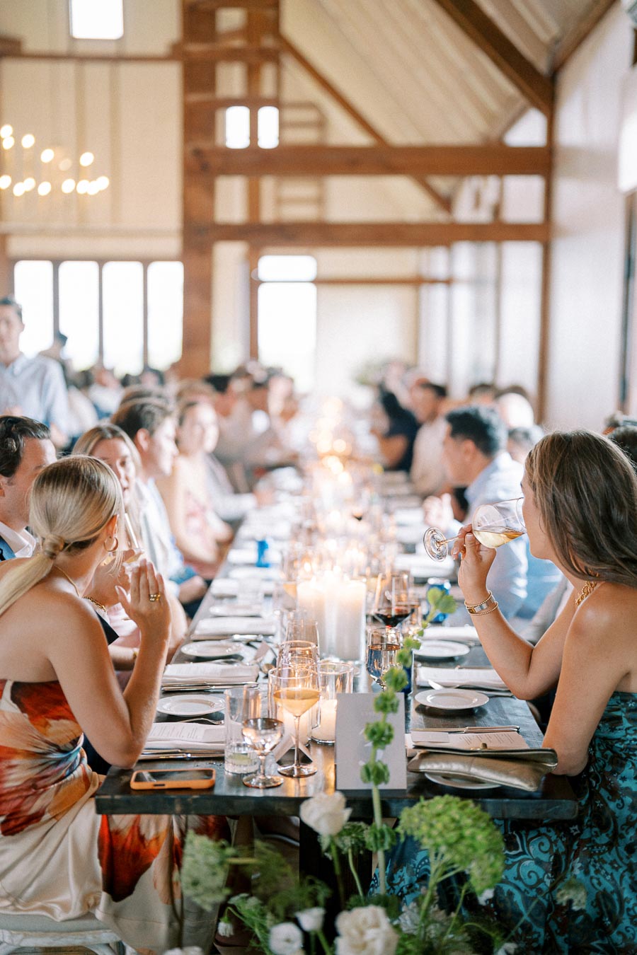 Elegant wedding reception in rustic venue with guests seated at a long table, adorned with candles and floral arrangements, creating a warm and intimate atmosphere.