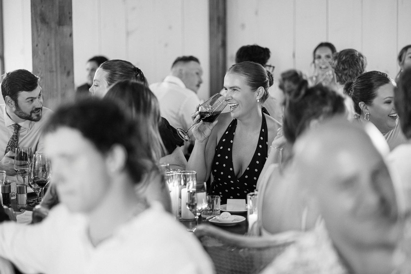 Black and white photo of a group of people enjoying a lively conversation at a social gathering, with a smiling woman in a polka dot dress holding a wine glass at the center.