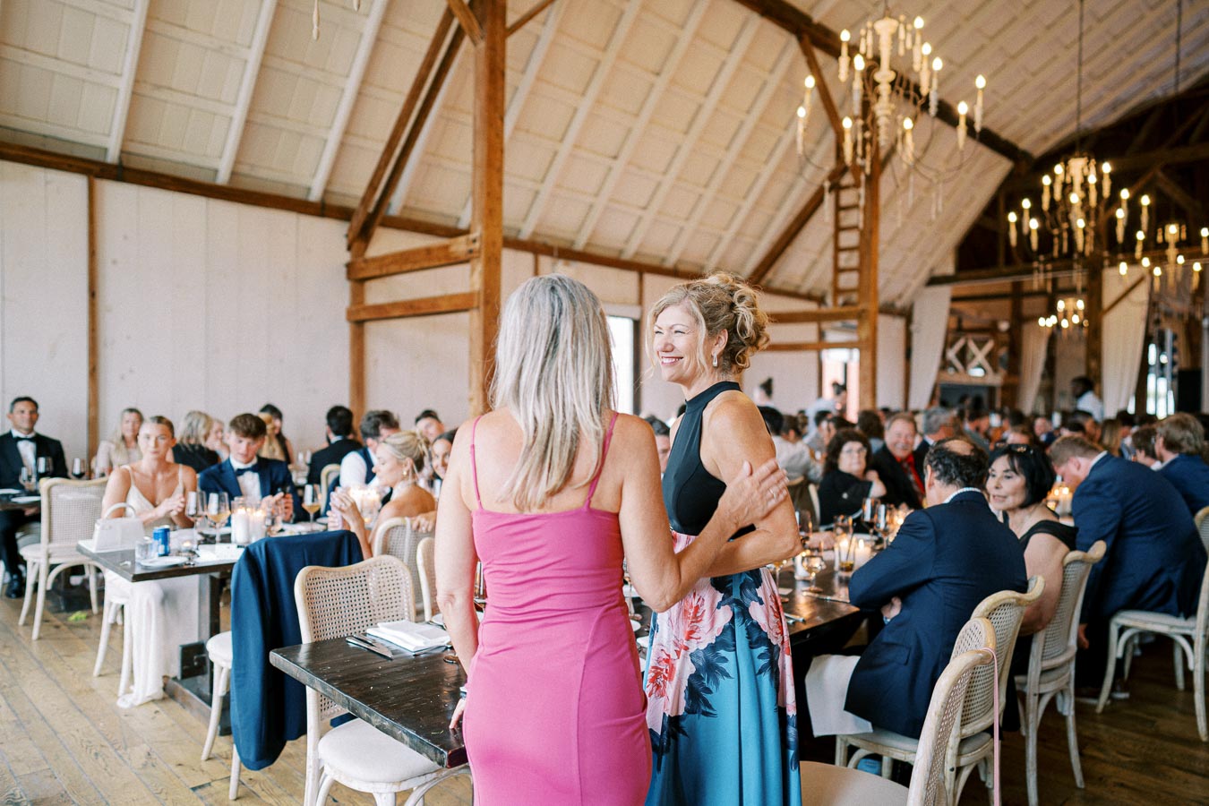 Elegant wedding reception in a rustic barn setting, featuring guests seated at decorated tables. Two women in vibrant dresses are standing and smiling, surrounded by elegantly dressed attendees and warm candlelight ambiance.