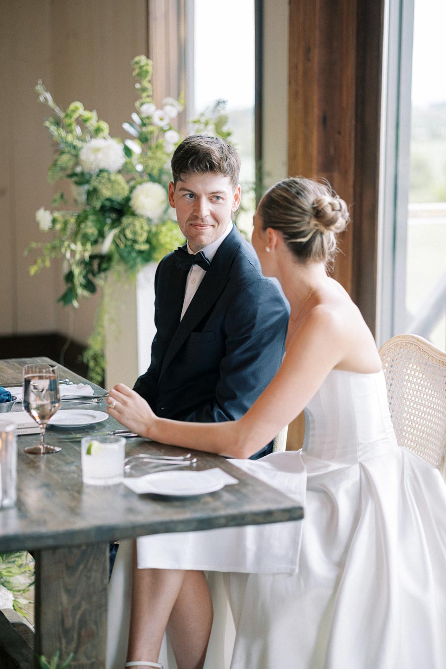 Elegant newlywed couple sitting at a beautifully decorated table during a wedding reception, with the bride in a stunning white gown and groom in a classic tuxedo, surrounded by lush floral arrangements and soft natural lighting.
