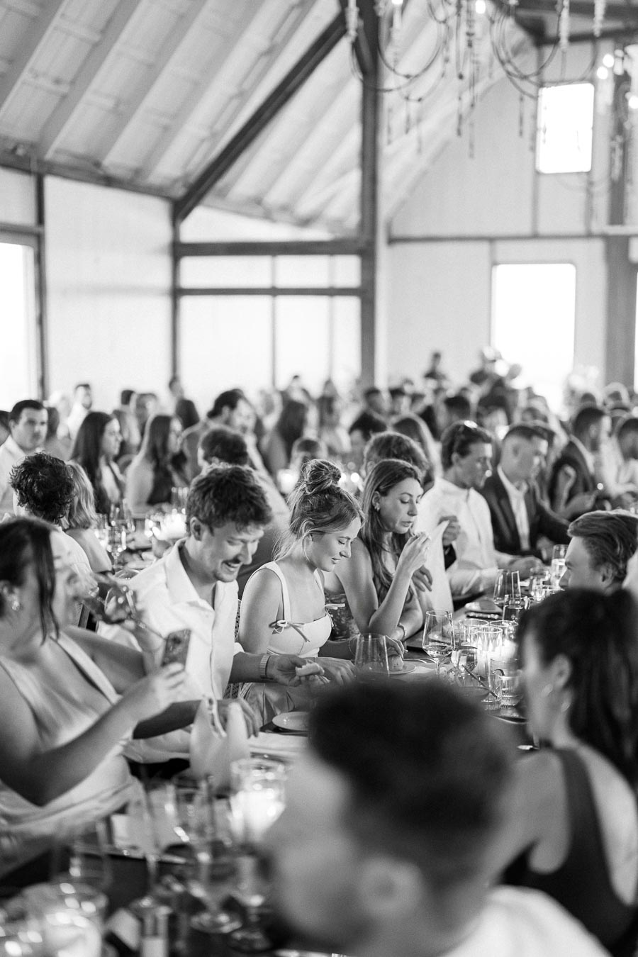 Black and white image of a lively gathering at a rustic indoor event venue, featuring guests seated at a long table, engaged in conversation and enjoying food and drinks under decorative lighting.