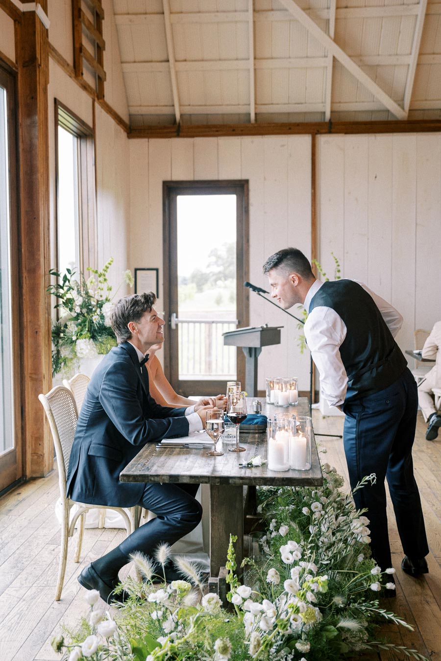 A man in a suit and bow tie sitting at a rustic wooden table, engaged in conversation with another man in formal attire in a bright, wooden room decorated with candles and flowers.