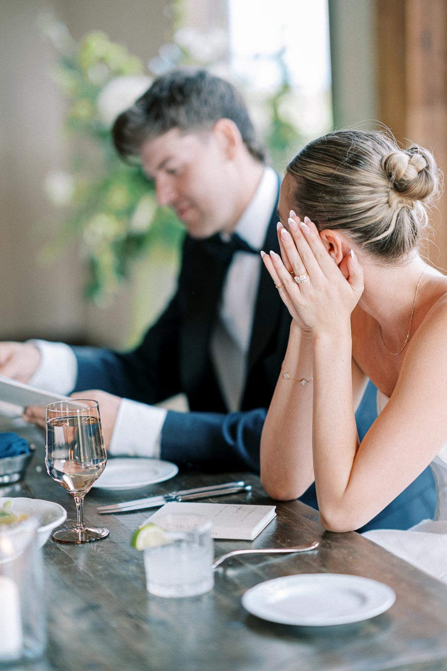 A couple dressed in formal attire seated at a table, with the woman playfully covering her face with her hands, highlighting her elegant engagement ring.