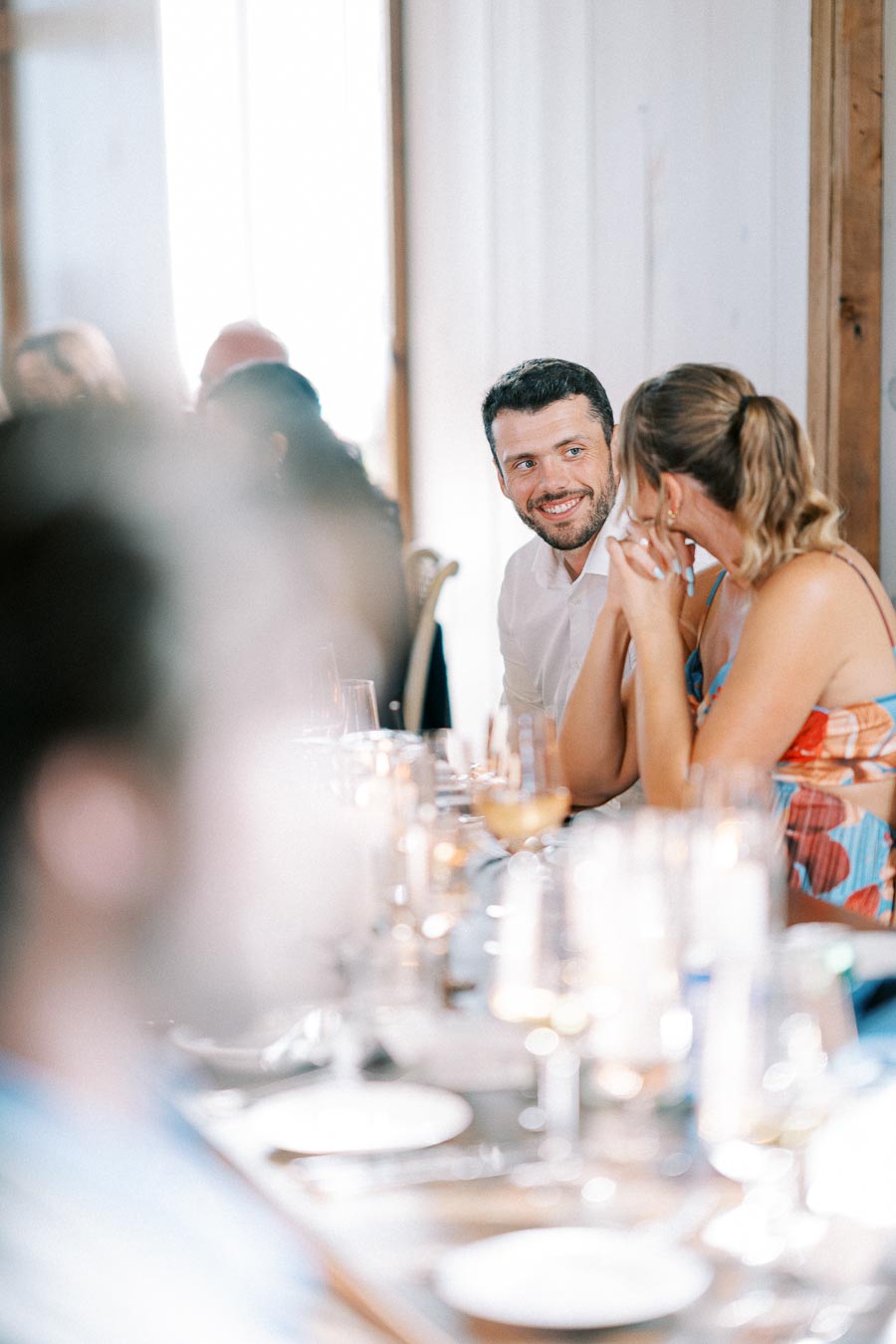 A joyful couple sitting at a decorated table during a wedding reception, surrounded by blurred guests, sharing a moment of laughter and conversation.
