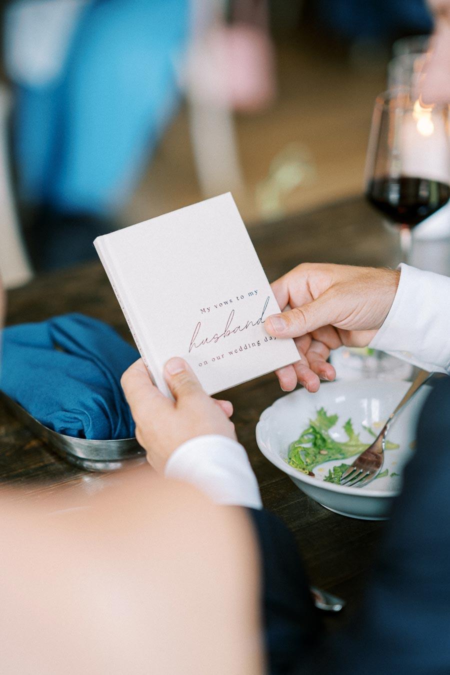A person holding a wedding vow booklet titled My vows to my husband on our wedding day at a dining table with a salad and a glass of red wine, symbolizing a romantic wedding celebration.