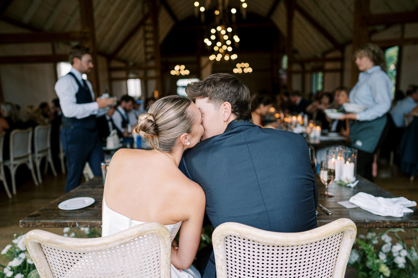 Couple sharing a tender moment at rustic wedding reception inside a beautifully decorated barn with chandeliers and candlelit tables.