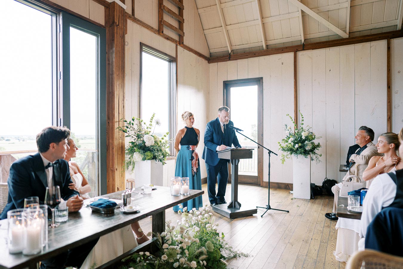 A man in a suit delivers a speech at a wedding reception, standing at a podium with a microphone in a bright, rustic venue. Guests, elegantly dressed, are seated attentively at tables adorned with candles and flowers, creating a serene and celebratory atmosphere.