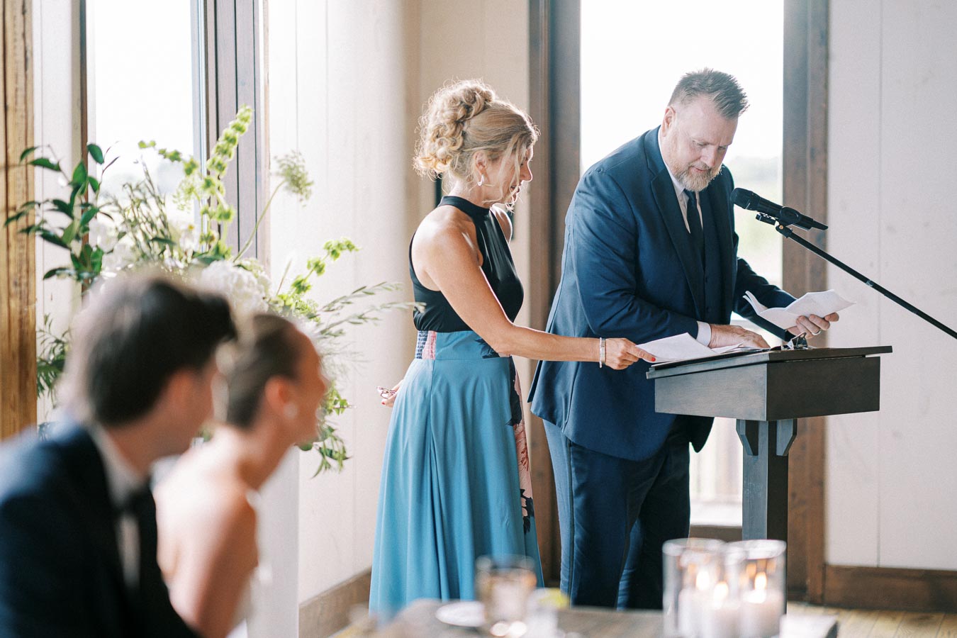 Guests giving a speech at a wedding reception, with a focus on a man and woman standing at a podium in formal attire, while surrounded by elegant floral arrangements and decor.