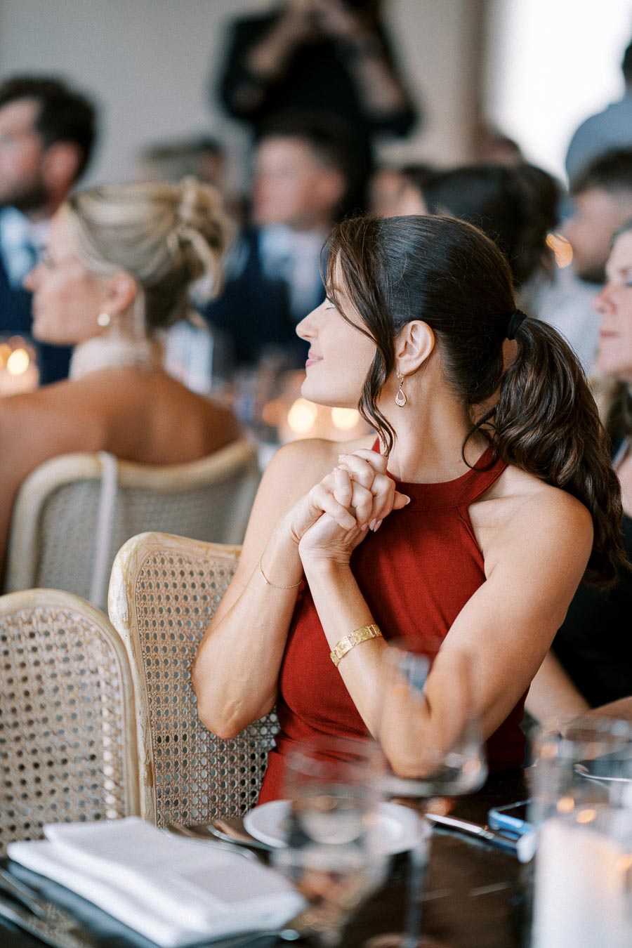 A woman in a red dress sitting at a formal event, looking thoughtful and smiling, surrounded by elegantly dressed guests in a softly lit setting.