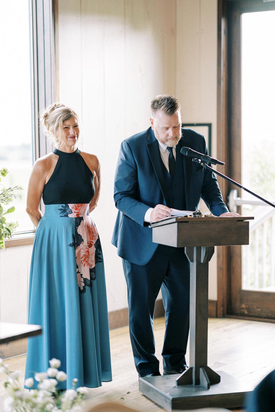Man in a navy suit reading at a podium next to a woman in a floral blue dress, inside a well-lit room with wooden floors and a window providing natural light.