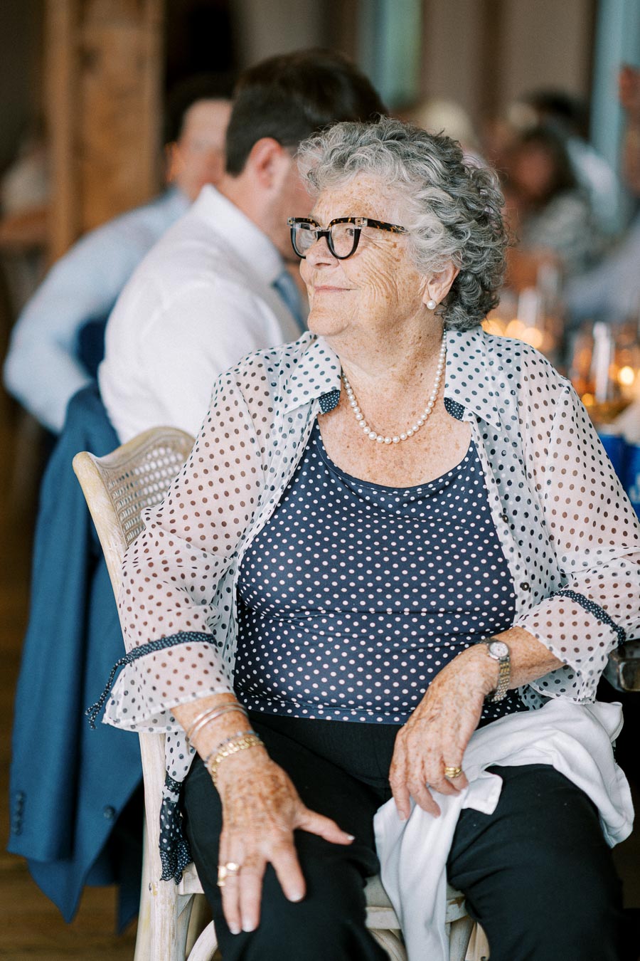 Elderly woman with glasses wearing a polka dot blouse and sheer jacket, seated at a social event.