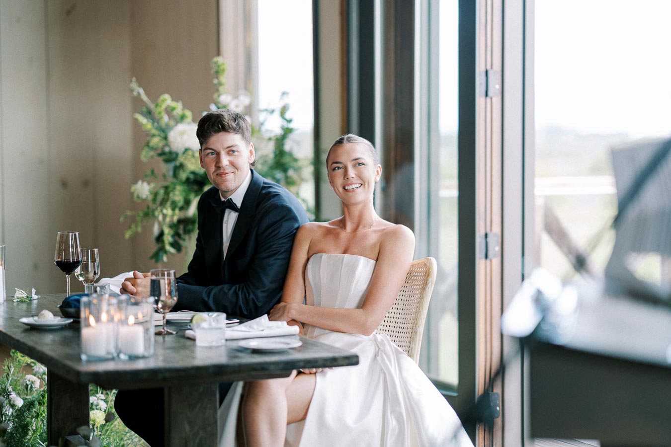 A smiling couple seated at a beautifully set wedding reception table by a large window. The groom is wearing a black suit with a bow tie, and the bride is in a strapless white gown. Decorative candles and flowers adorn the table, with greenery visible in the background.