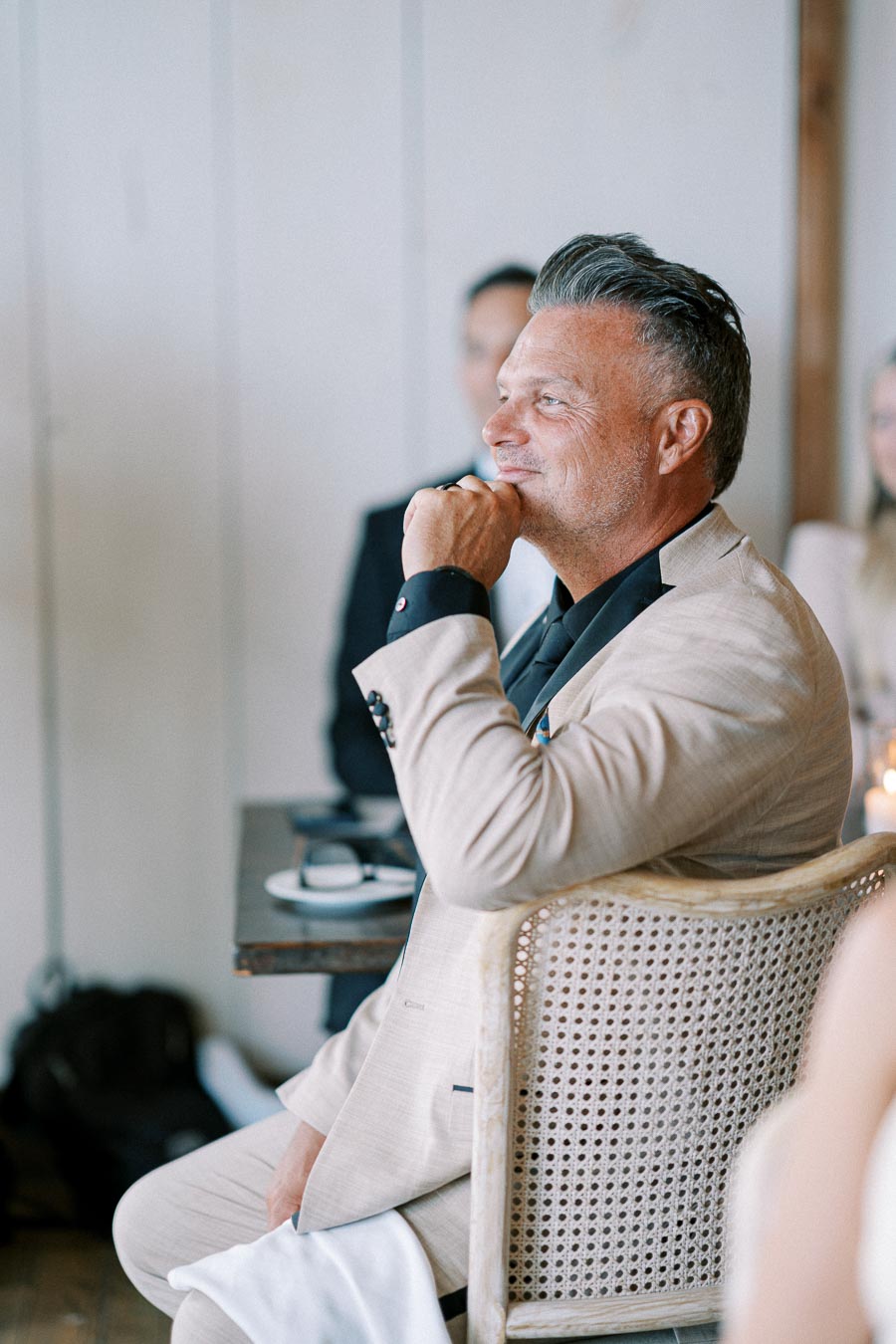A man in a beige suit sitting on a wicker chair, smiling thoughtfully during an indoor event.