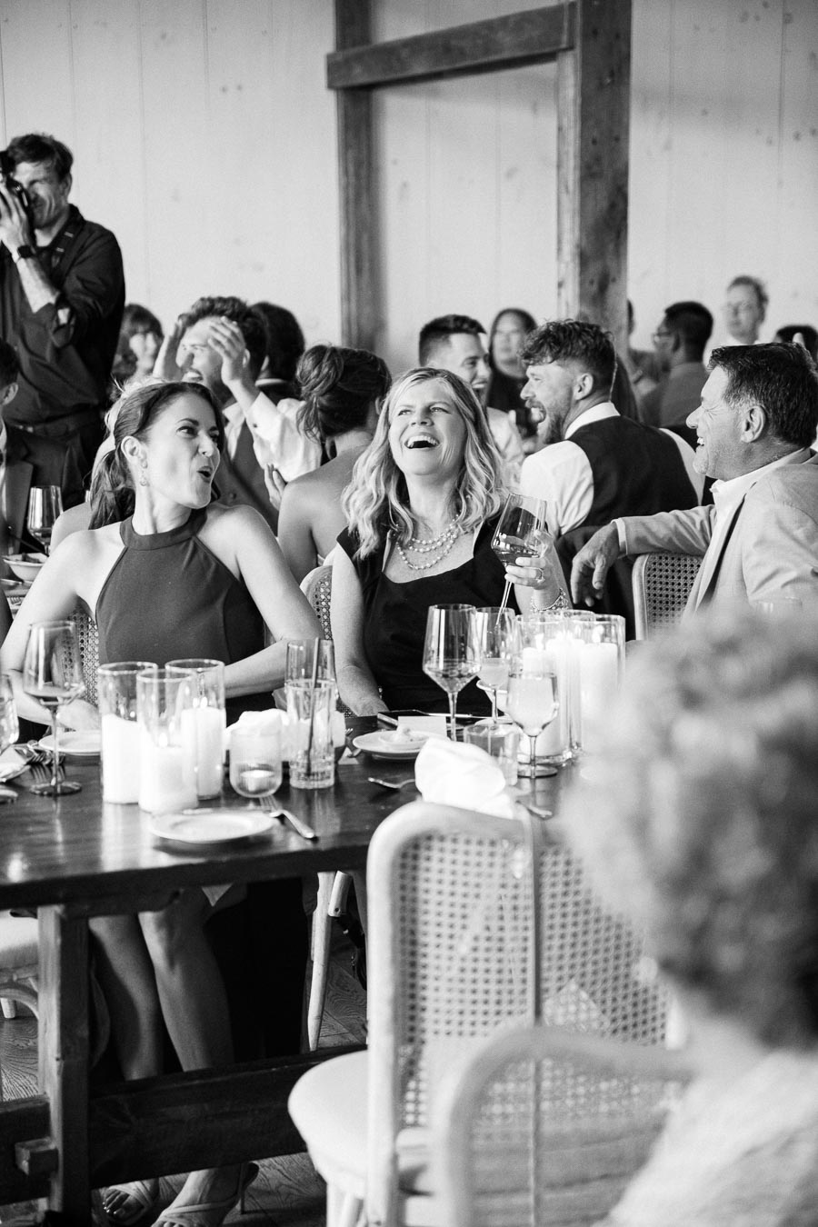 Black and white photo of a lively wedding reception with guests laughing and enjoying themselves at a beautifully set table, enhancing the joyful atmosphere.