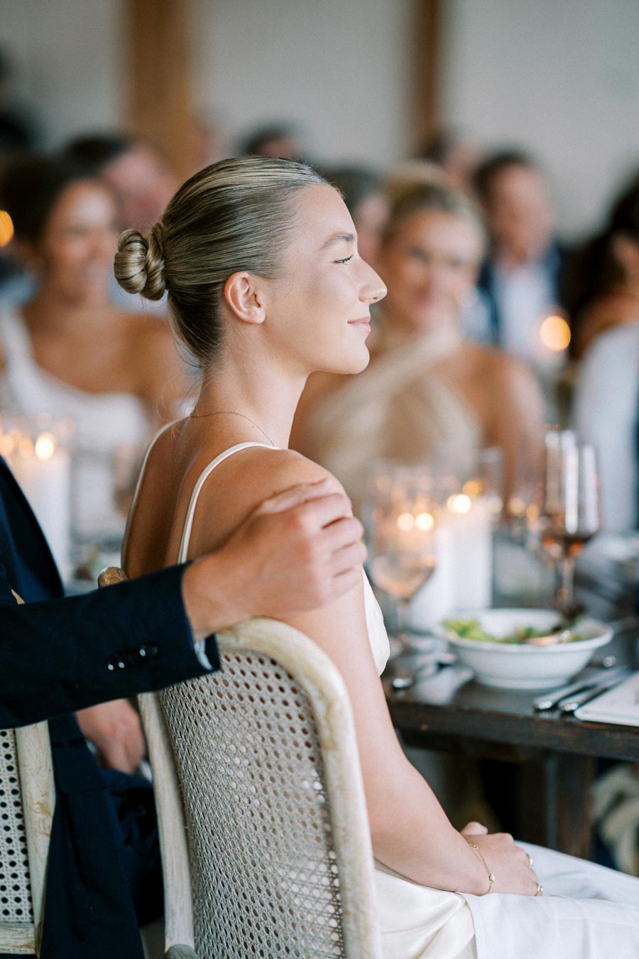 Bride sitting at a wedding reception, eyes closed and smiling, with a hand gently resting on her shoulder, surrounded by elegant table settings and warmly lit candles.