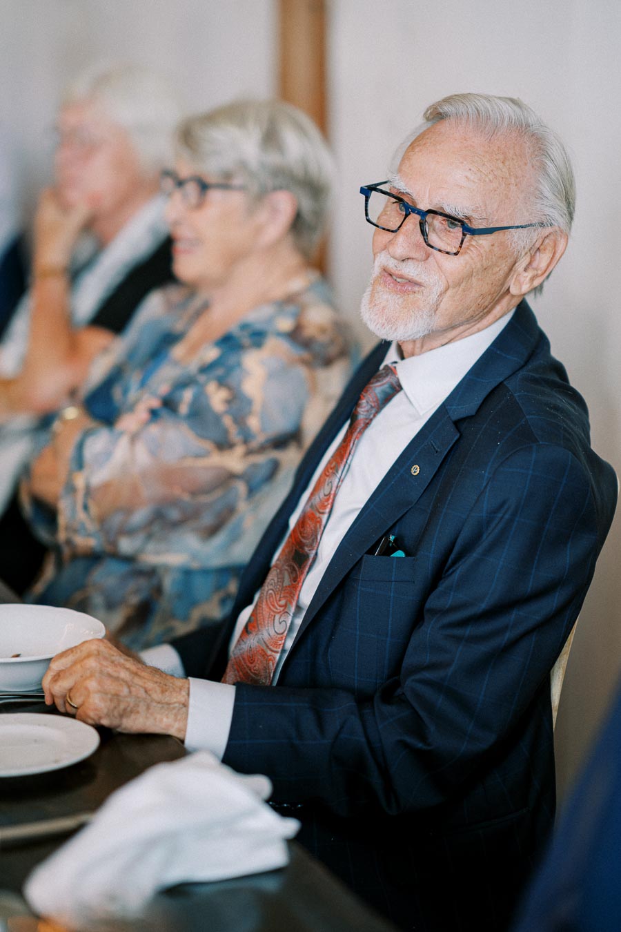 Elderly man in glasses wearing a suit and tie sits at a table during an event, with people in the background.