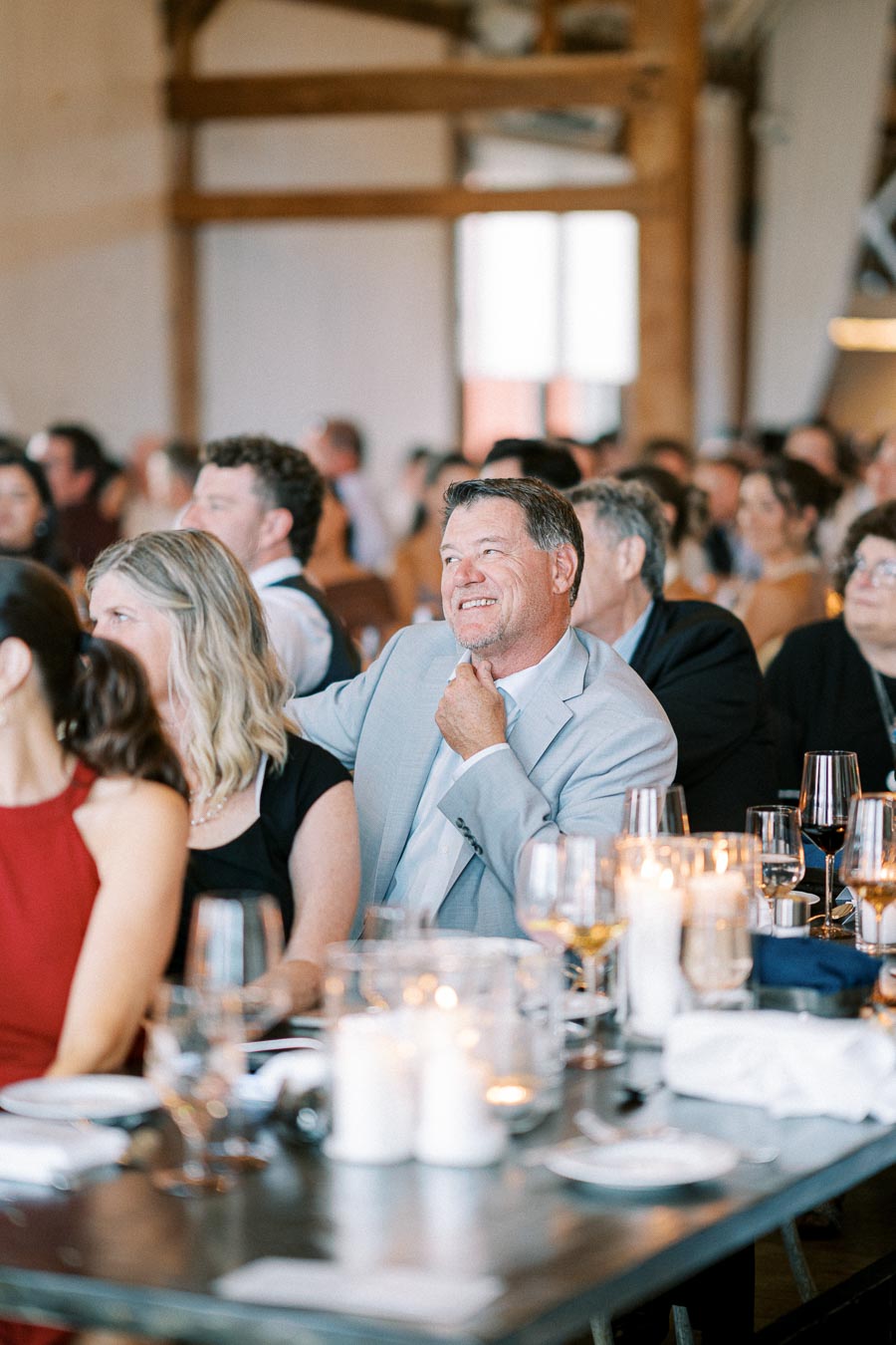 Guests enjoying a joyful moment at a wedding reception, seated around candlelit tables with wine glasses in a rustic venue.