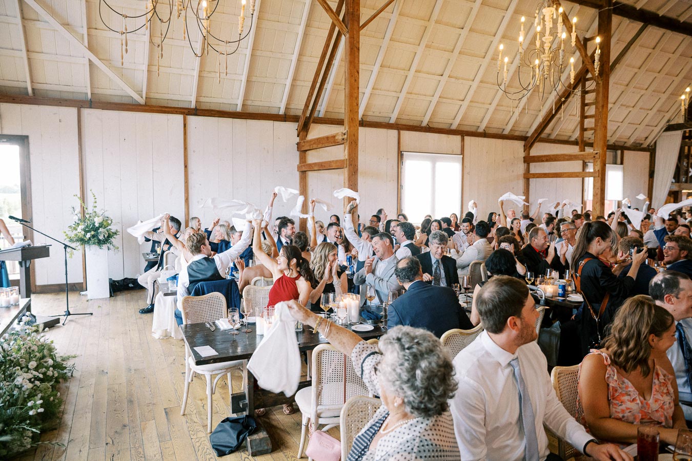 A joyous wedding reception inside a rustic barn venue, with guests enthusiastically waving napkins and seated at elegantly decorated tables.