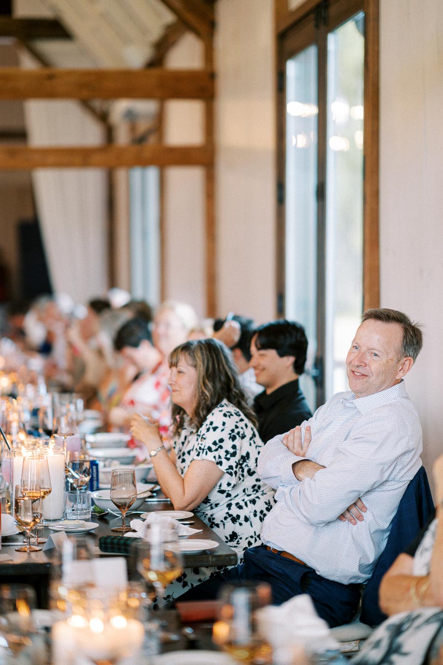 Guests enjoy a serene dining experience at a warmly lit event, featuring a long table adorned with candles and elegant glassware in a rustic setting.