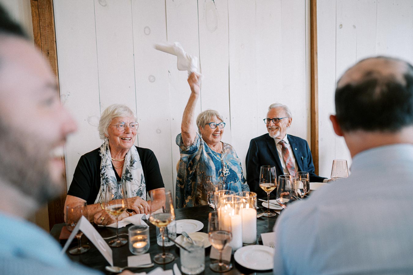 Elderly people enjoying a celebratory dinner, with a woman joyfully raising a napkin, surrounded by friends and candlelit table settings.