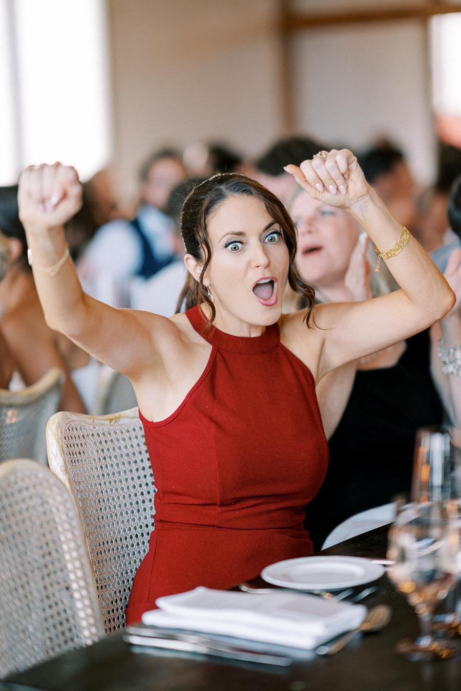 A woman in a red dress appears excited and surprised, raising her arms while seated at a formal event with other attendees in the background.