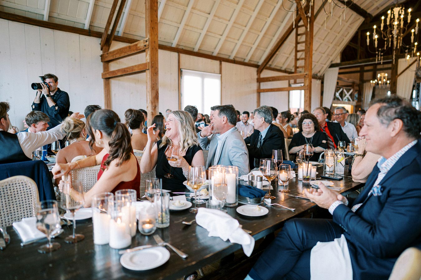 A lively wedding reception with guests laughing and enjoying themselves in a rustic barn setting, candles and wine glasses adorn the table, and a photographer capturing moments.
