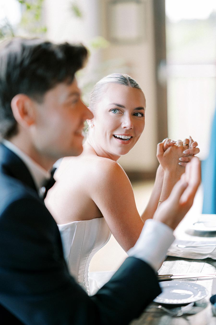 Wedding reception scene with a smiling bride in a white dress, enjoying a conversation at a table, set with plates and silverware, alongside a blurred male guest in a tuxedo.