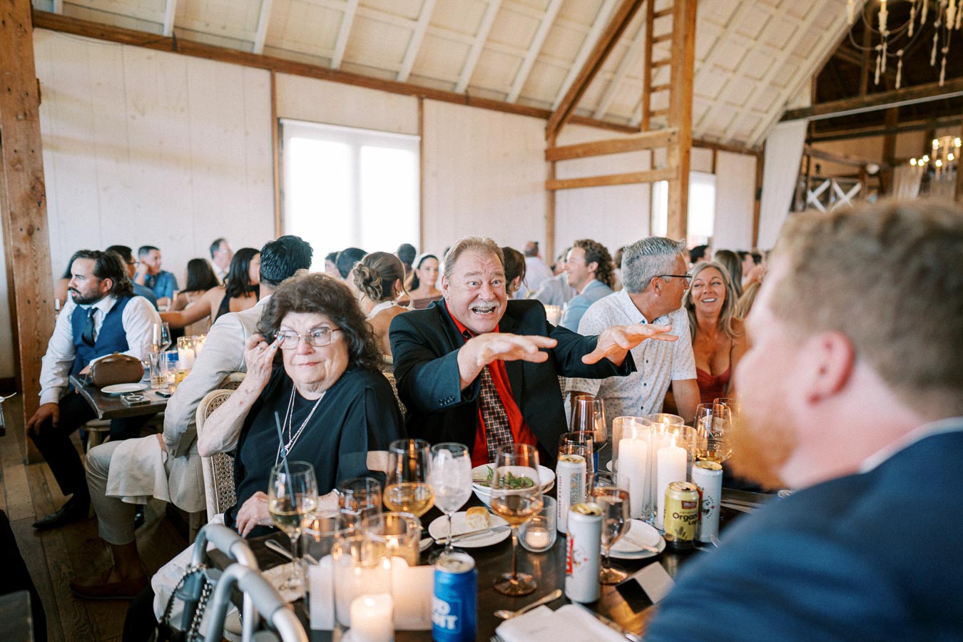 A festive group of people enjoying a banquet inside a rustic venue, with tables adorned with elegant candles and beverages. A cheerful man in a suit playfully gestures across the table, creating a lively and joyful atmosphere.