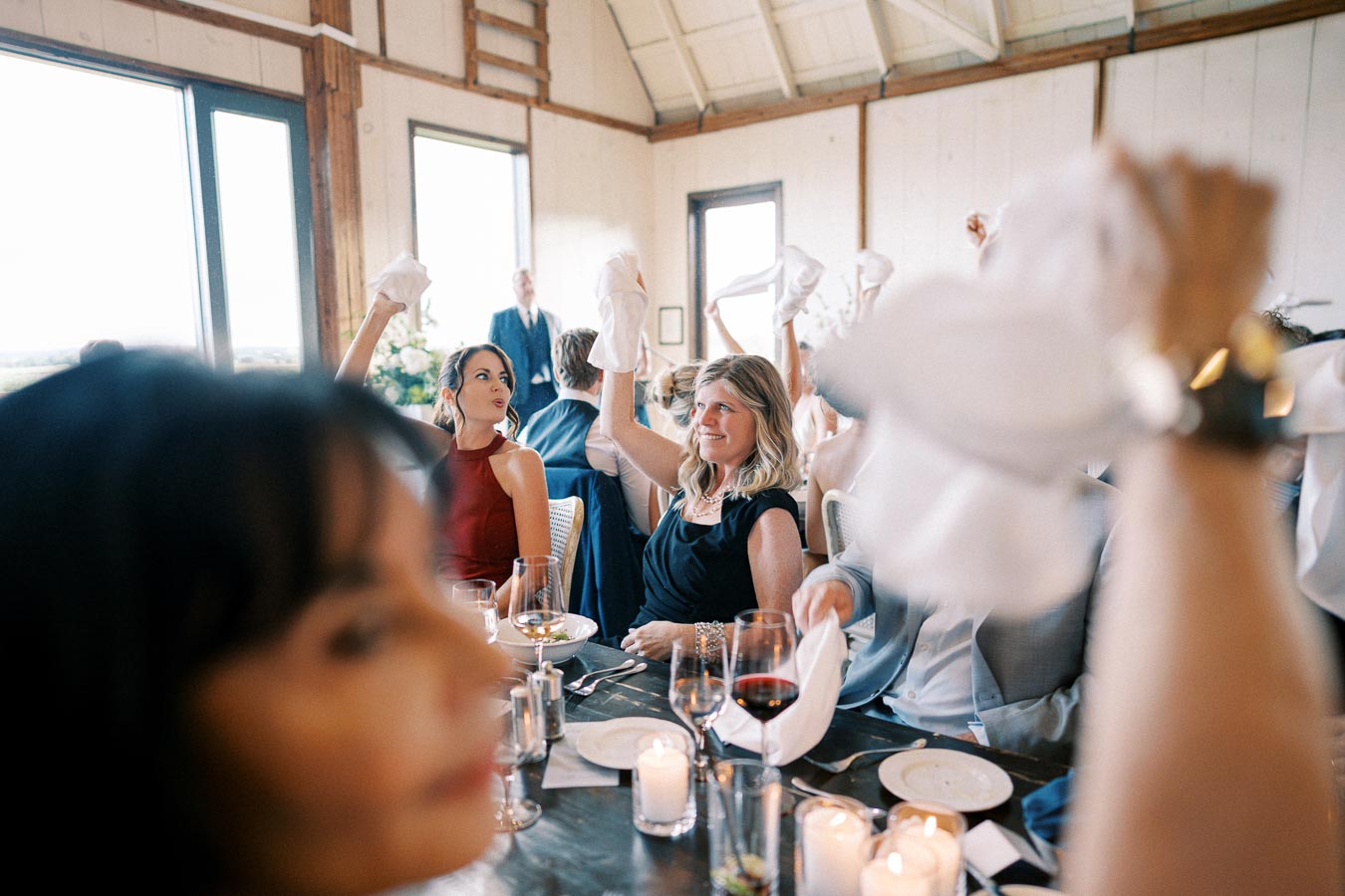 Guests waving napkins in celebration during a wedding reception inside a rustic venue, with glasses of wine and candles on the table, creating a festive and joyful atmosphere.