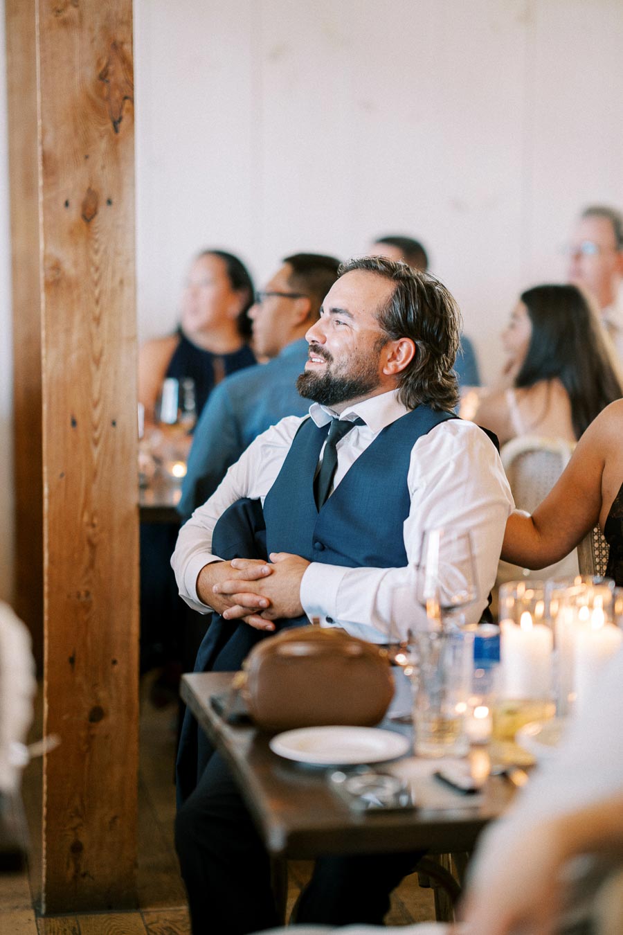 Man in a formal blue vest and tie smiling during an event, surrounded by attendees at a candlelit dinner gathering.