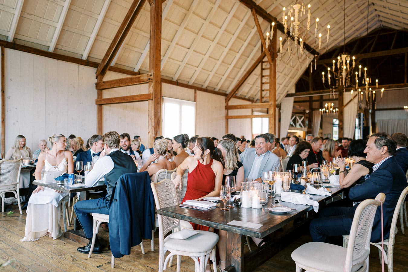 A lively wedding reception in a beautifully decorated barn setting, featuring elegantly dressed guests seated at rustic wooden tables adorned with candles and wine glasses, under a ceiling with chandeliers.
