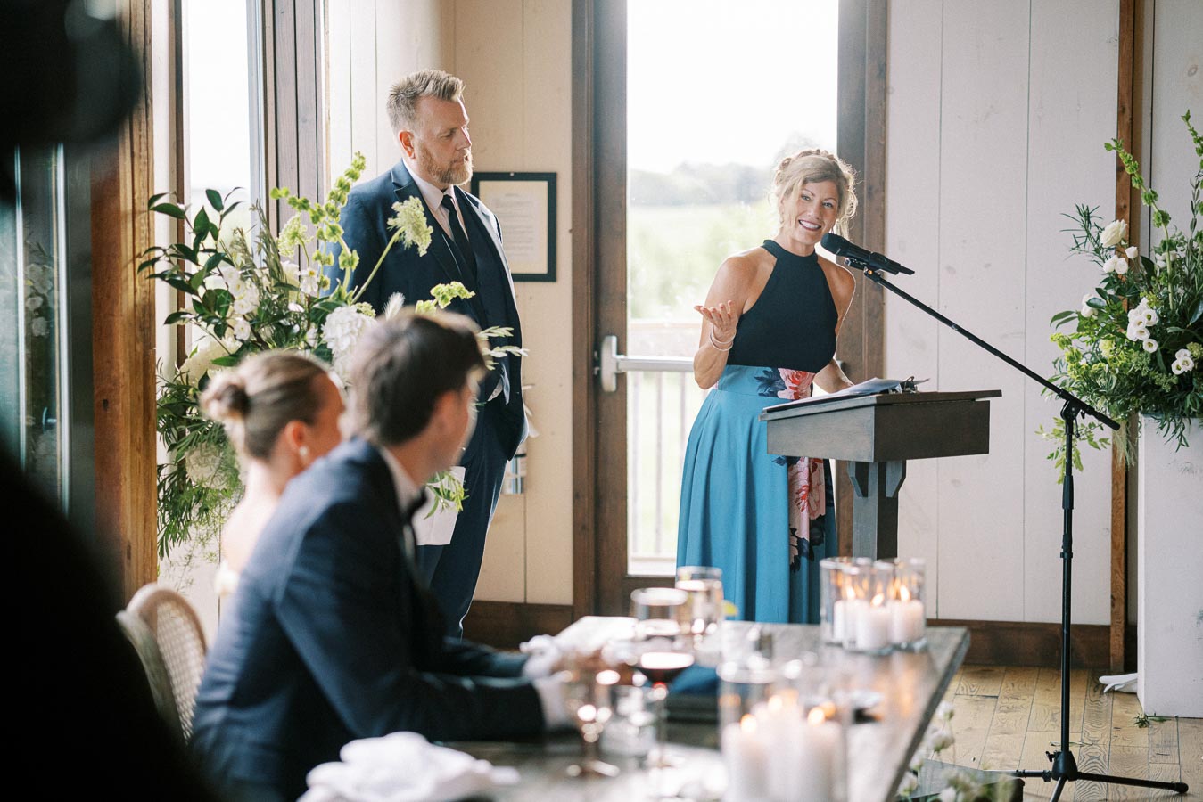 A woman in a blue dress delivers a speech at a wedding reception, standing at a podium with a microphone. A man in a suit stands beside her, and guests seated at a table are listening. The room features large windows, floral arrangements, and lit candles on the table, creating an elegant and festive atmosphere.