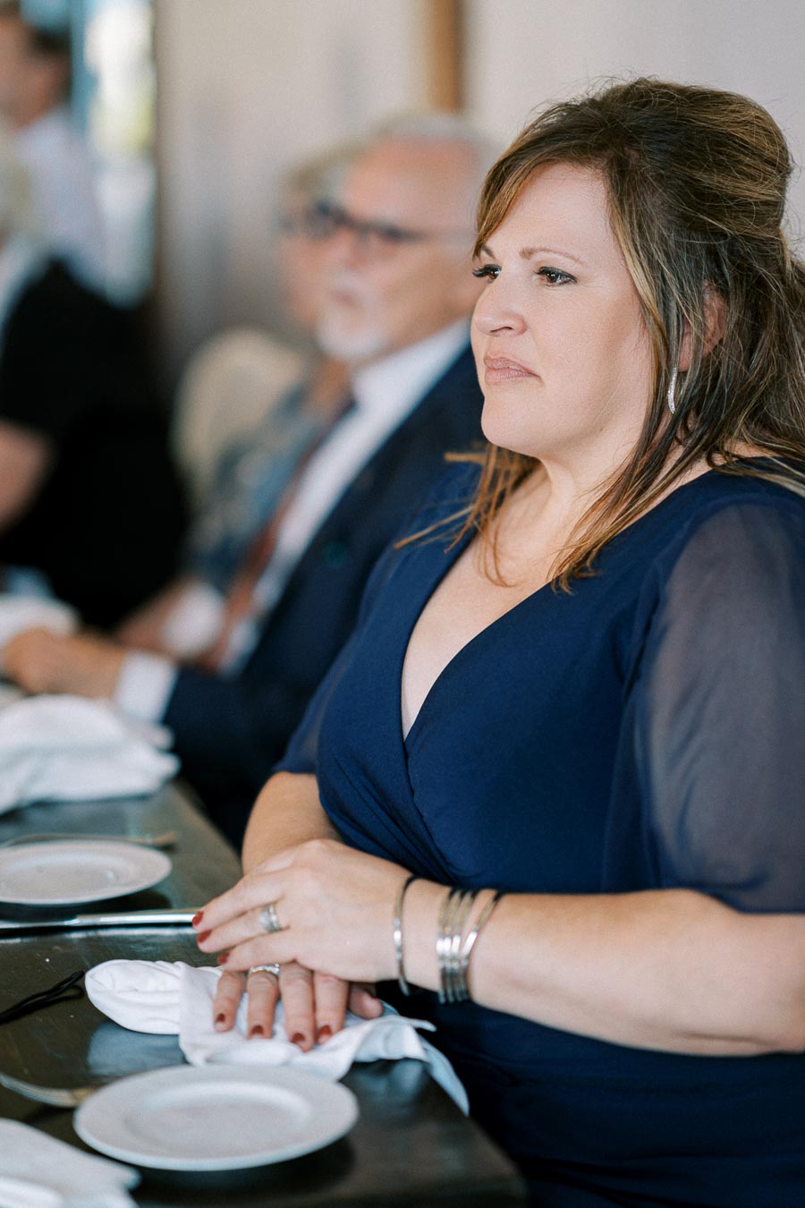 Woman in a blue dress seated attentively at a formal event, with a blurred background of other attendees and table settings.