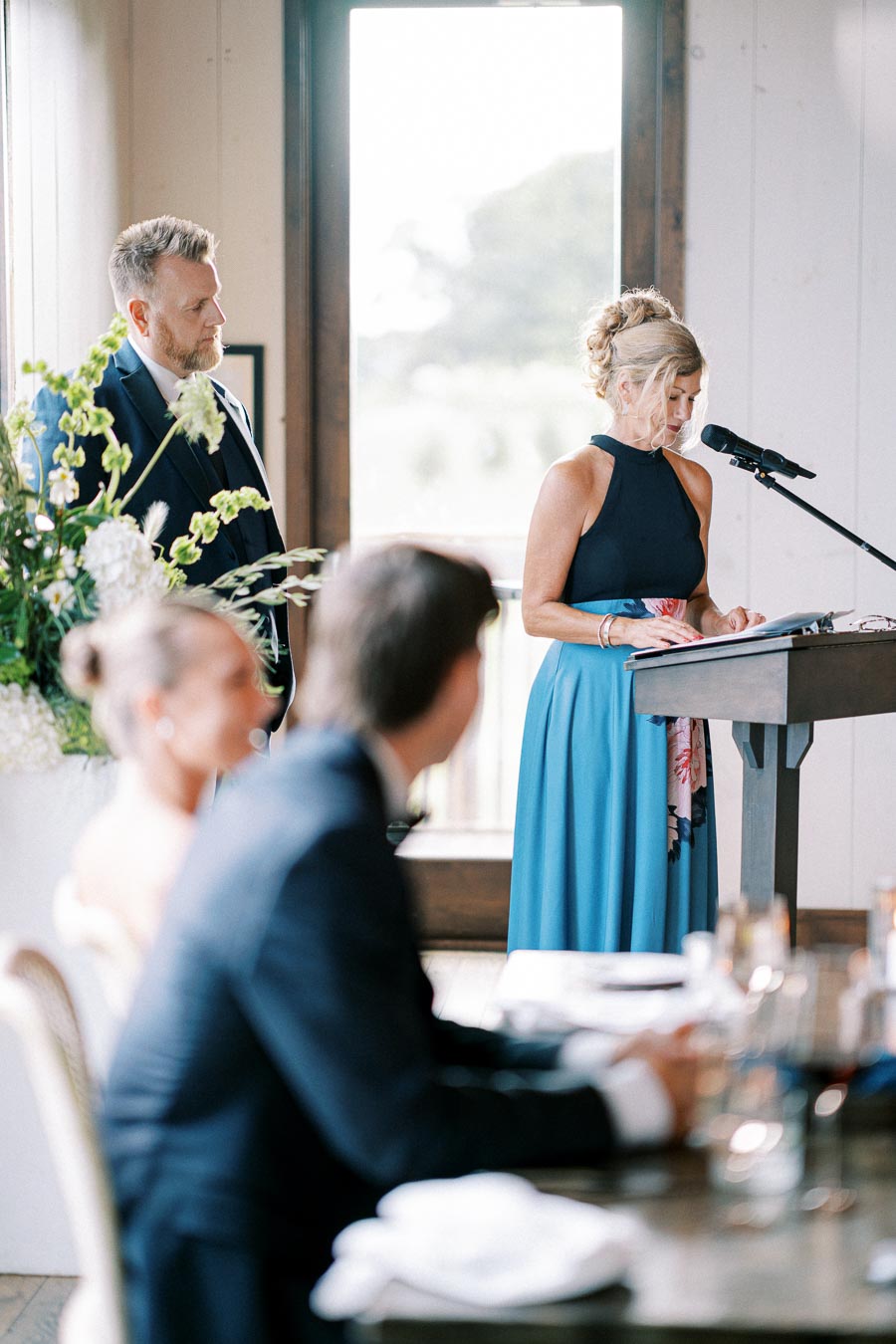 A woman in a blue dress delivers a speech at a wedding reception, standing at a podium with a microphone. A man in a suit stands nearby listening, while guests sit at a table in the foreground with floral decorations.