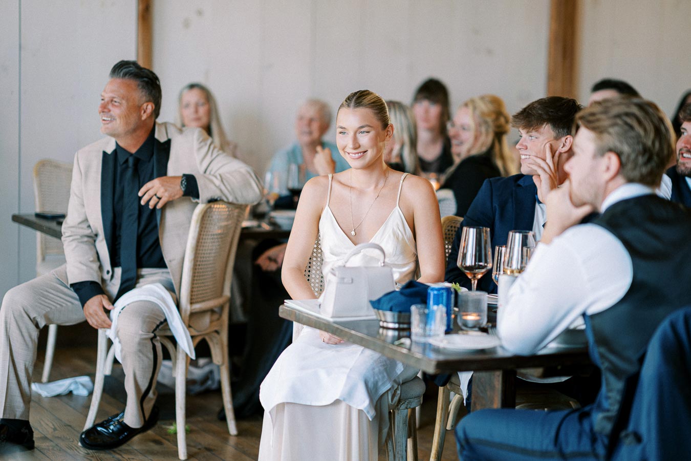 A cheerful group of well-dressed guests enjoying an elegant indoor event, seated at tables with wine glasses and decor in a stylish venue.