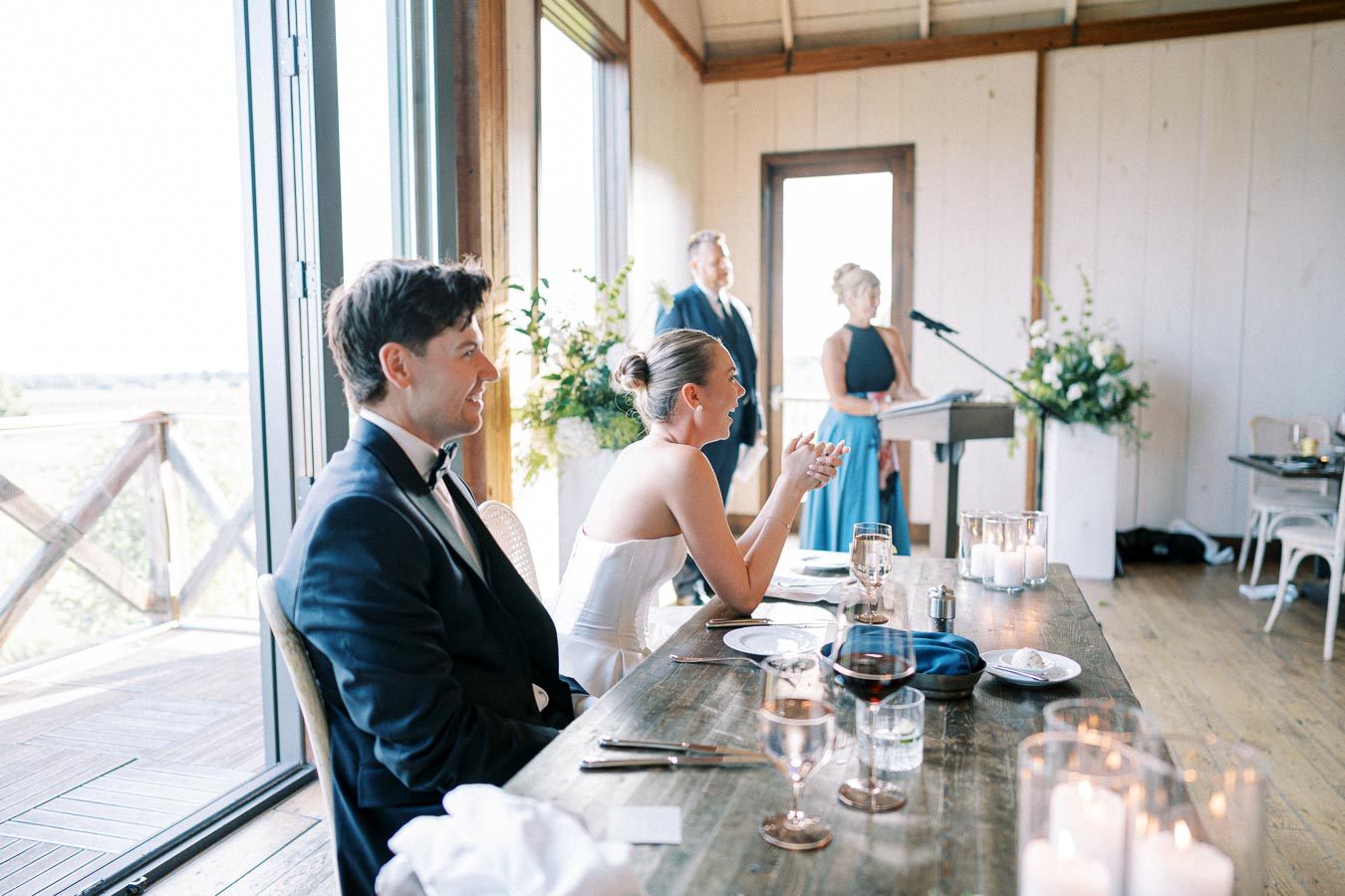 Bride and groom laughing during wedding reception speech inside a beautifully decorated venue with large windows and candlelit tables.