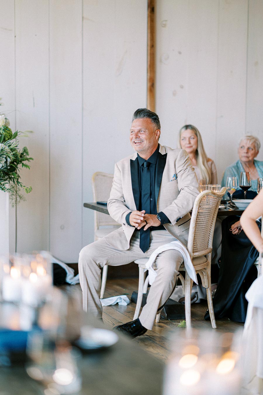 A man in a beige suit sits smiling at a table during an indoor event, surrounded by elegantly dressed guests, with candles and greenery in the decor.