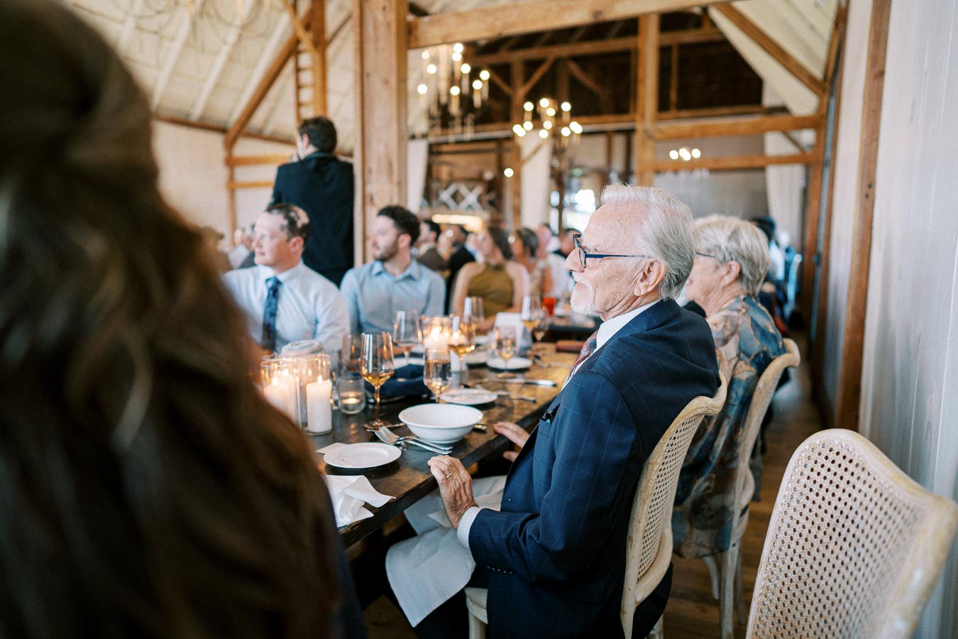 A group of elegantly dressed guests seated at a wooden table during a wedding reception in a rustic barn setting, featuring lit candles and decorative chandeliers.
