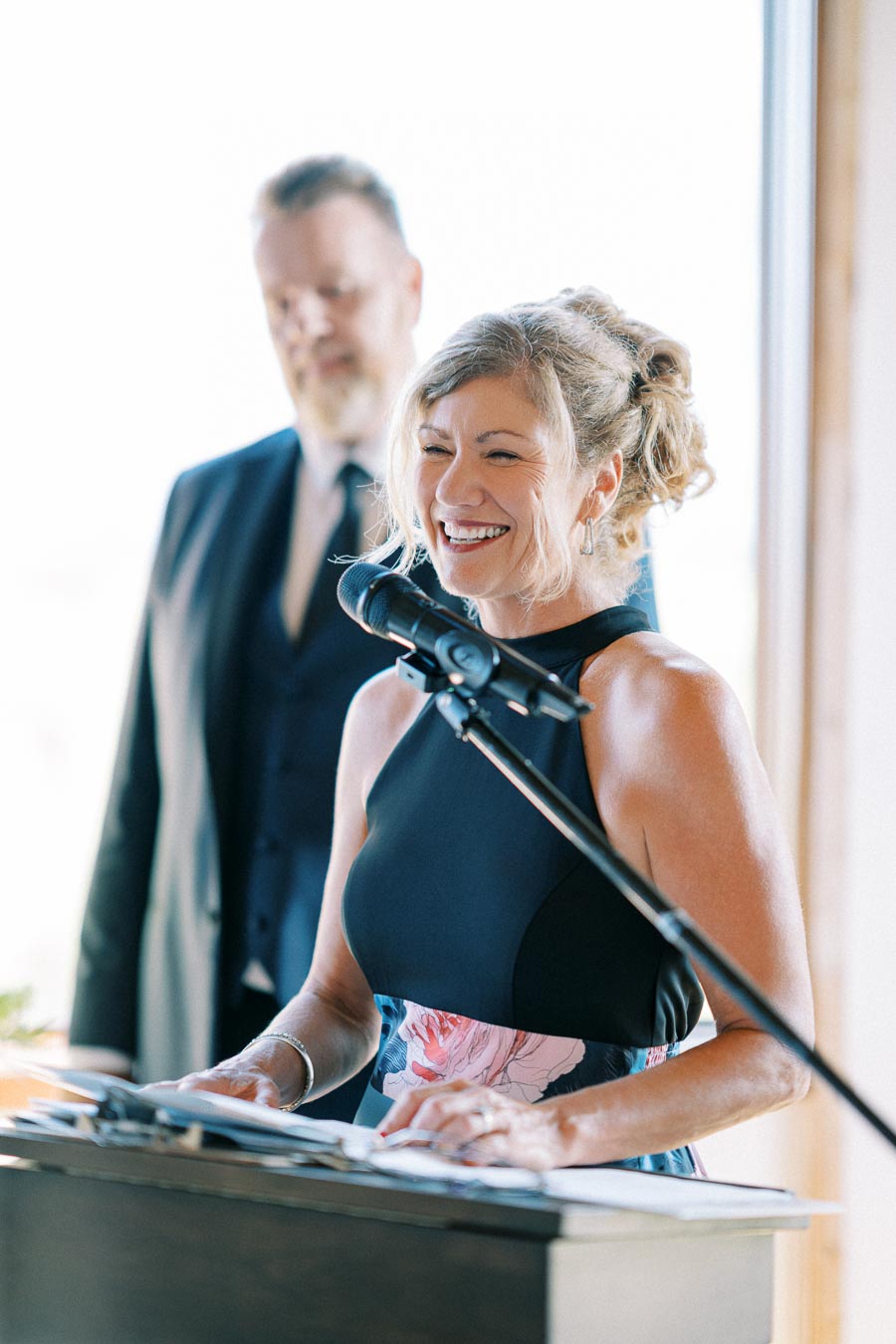 A woman in a floral dress smiles while speaking into a microphone at a podium, with a man in a suit standing in the background near a bright window.