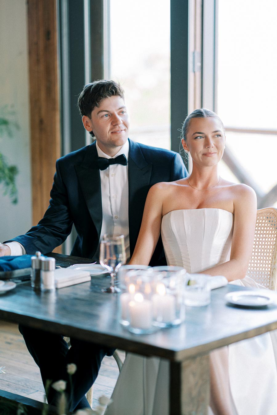 Elegant couple at wedding reception, groom in black tuxedo and bride in strapless white gown, seated at candlelit table indoors.