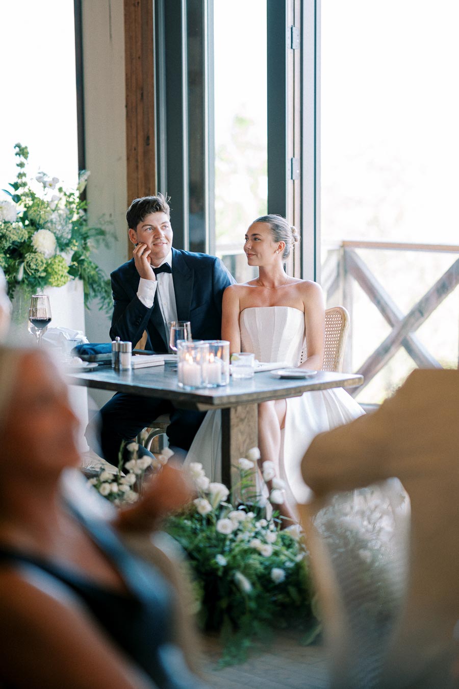 A beautifully dressed wedding couple sits at a decorated table during a reception, surrounded by lush floral arrangements and soft candlelight, creating a romantic ambiance.