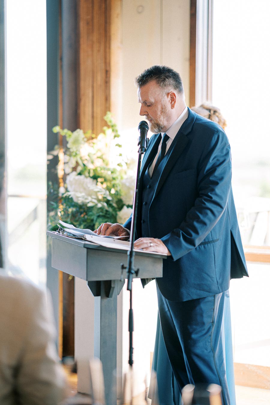 A man in a blue suit giving a speech at a podium with a microphone, in a bright room with floral decorations in the background.