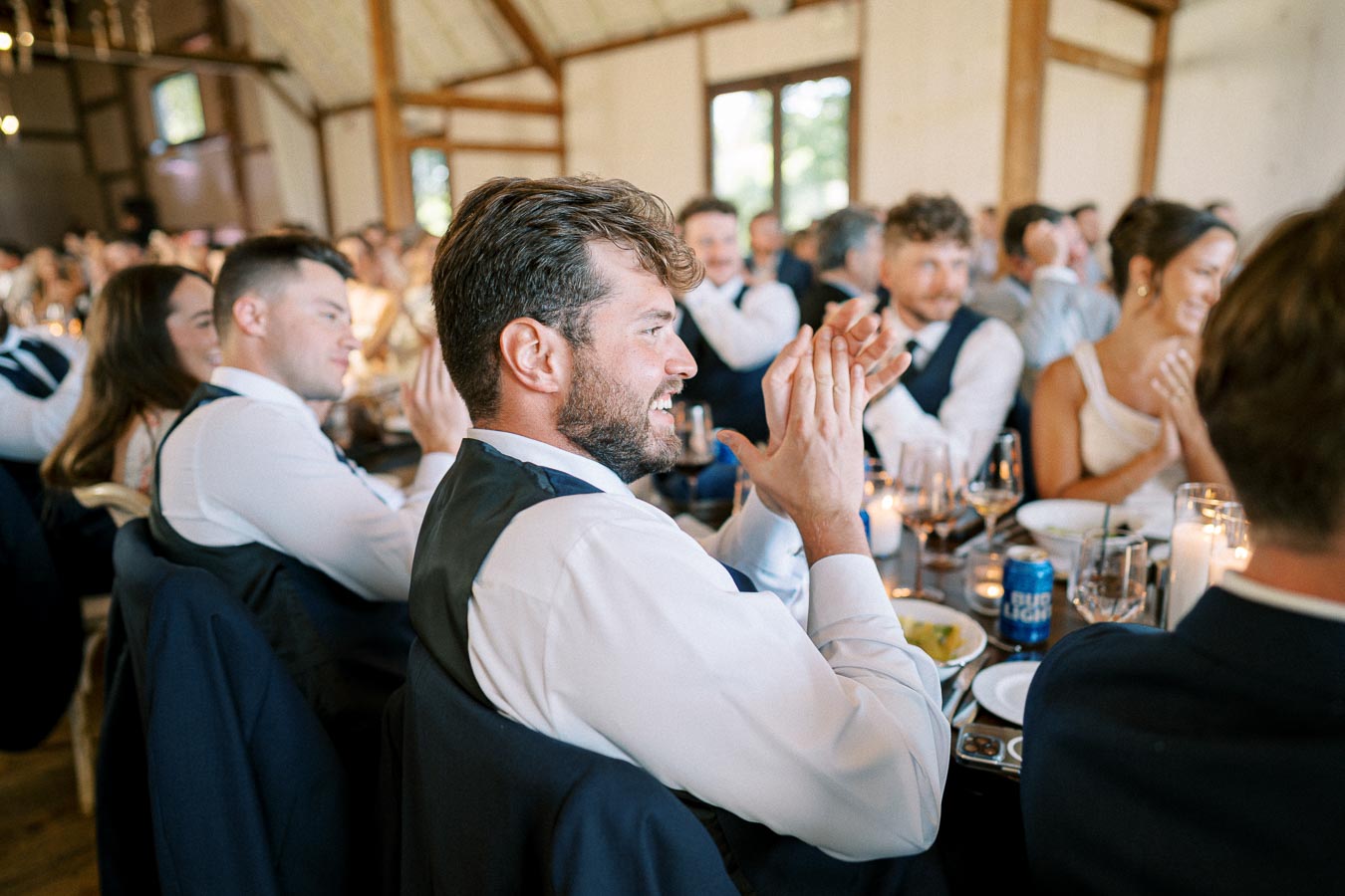 A joyful man in a suit clapping at a lively wedding reception, surrounded by guests sitting at elegantly set tables with glasses and candles, creating a warm and celebratory atmosphere.