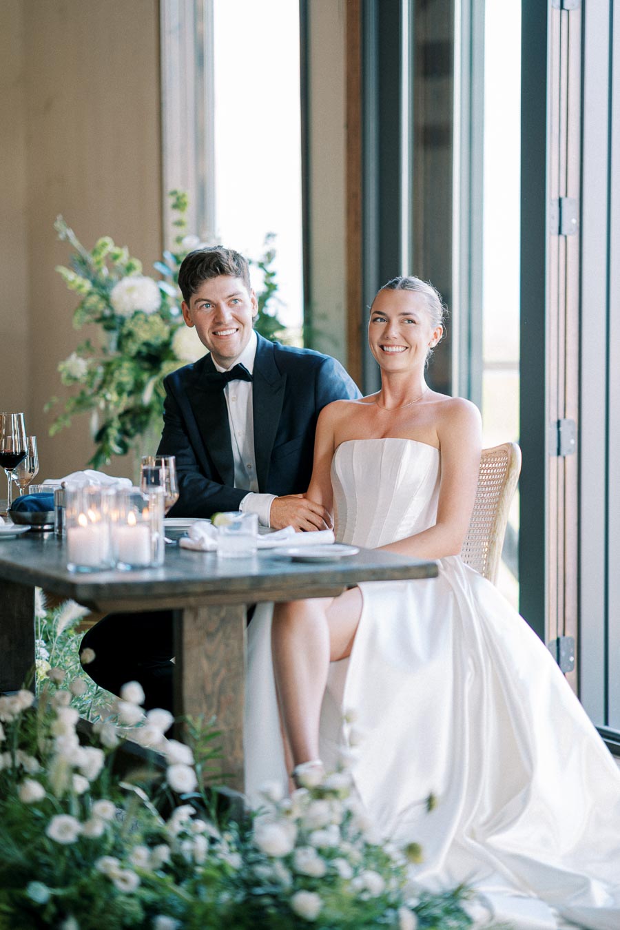 Smiling bride and groom seated at a beautifully decorated wedding table with floral arrangements and candles, capturing a joyful moment during their celebration.