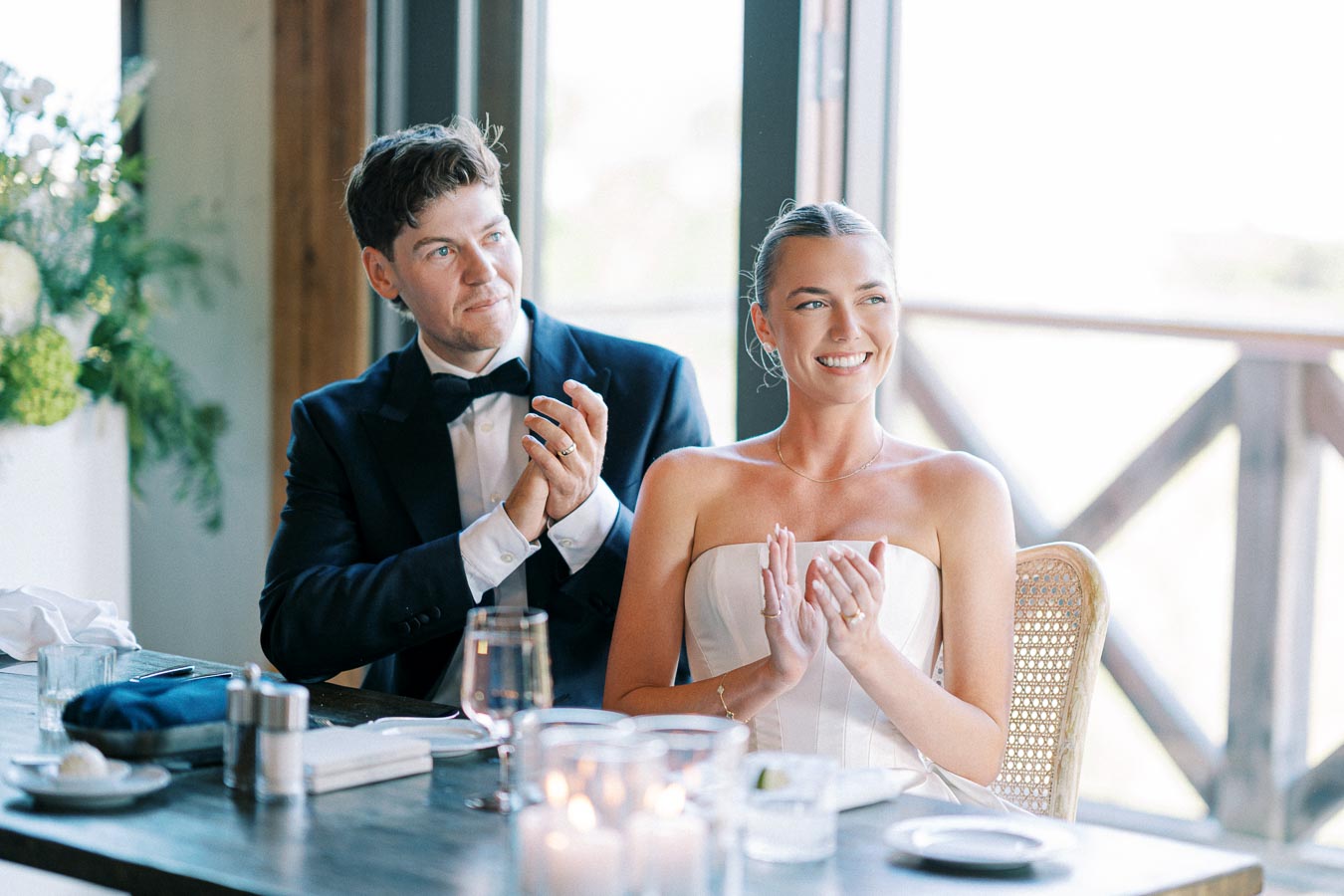 Wedding celebration with smiling bride in a strapless dress and groom in a tuxedo clapping hands at a dining table, illuminated by natural light from large windows.