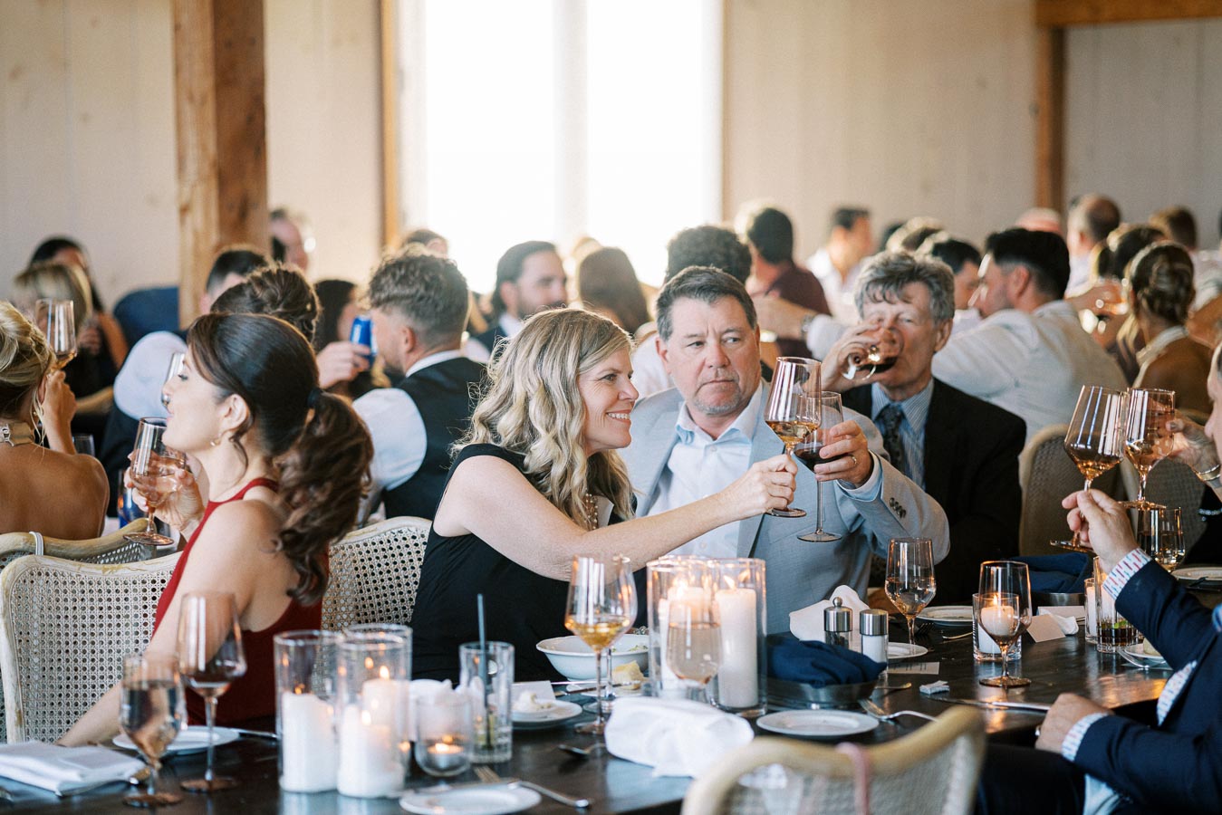 Group of people raising their glasses in a toast at an elegant indoor event, with candles and table settings creating a warm, celebratory atmosphere.
