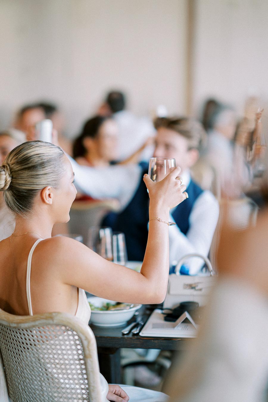 Elegant woman in a white dress raising a glass in a celebratory toast at a formal gathering, with blurred guests in the background.